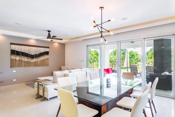 Modern open-plan living and dining room with cream seating, dining table, macramé wall art, and sliding glass doors opening to tropical garden patio.