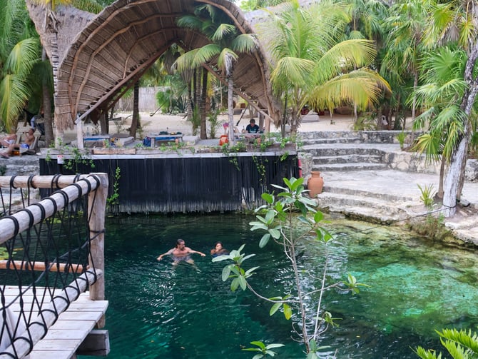 Two swimmers in a turquoise cenote with a wooden bridge and palm trees