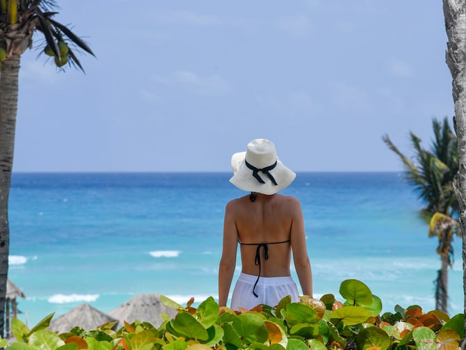 Woman in a sun hat looking out at turquoise Caribbean water through palm trees