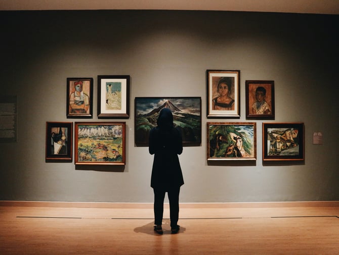 Person standing before a wall of paintings in a warm gallery