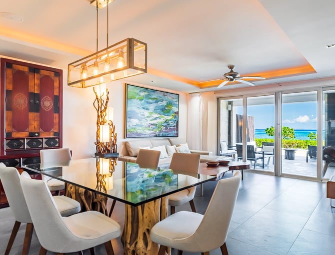 Modern dining room with glass table, cream chairs, ocean view through floor-to-ceiling doors, and red lacquered cabinet.